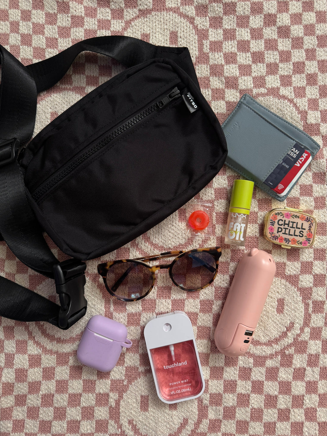 Black crossbody plus-size bag. The contents of the bag are displayed next to the bag, sunglasses, mints, wallet, pill case, pepper spray, and lip balm. Over a smiley checkered backdrop. 