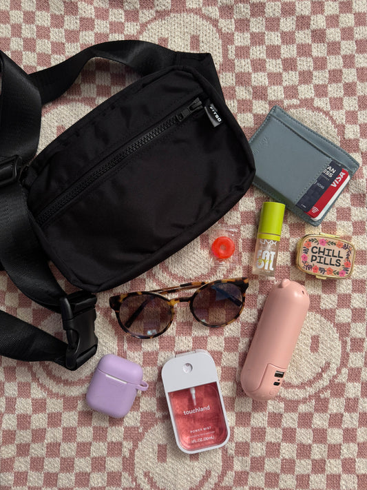 Black crossbody plus-size bag. The contents of the bag are displayed next to the bag, sunglasses, mints, wallet, pill case, pepper spray, and lip balm. Over a smiley checkered backdrop. 
