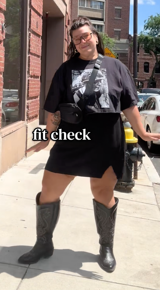 plus size woman wearing a black graphic tee crop top, a black, skirt, black western boots with a black crossbody bag accessory on the streets of Boston. 