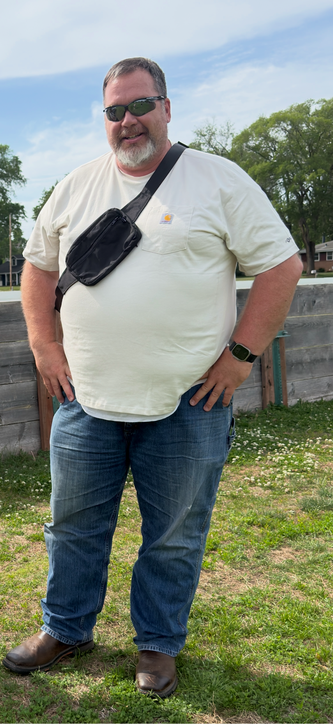 Man wearing sunglasses and a white t-shirt with a black belt bag over his shoulder, standing outdoors.